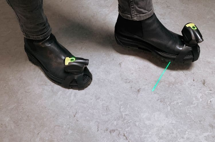 A photo of the Path Finder device strapped on to a pair of black boots. The green laser shines on the ground.