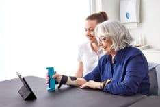 A photo of a woman with a bandage on her right wrist using the GripAble device, sitting besides a physiotherapist.