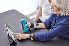 A photo of a woman with a bandage on her right arm using the GripAble device and pointing at a tablet sitting on a table.