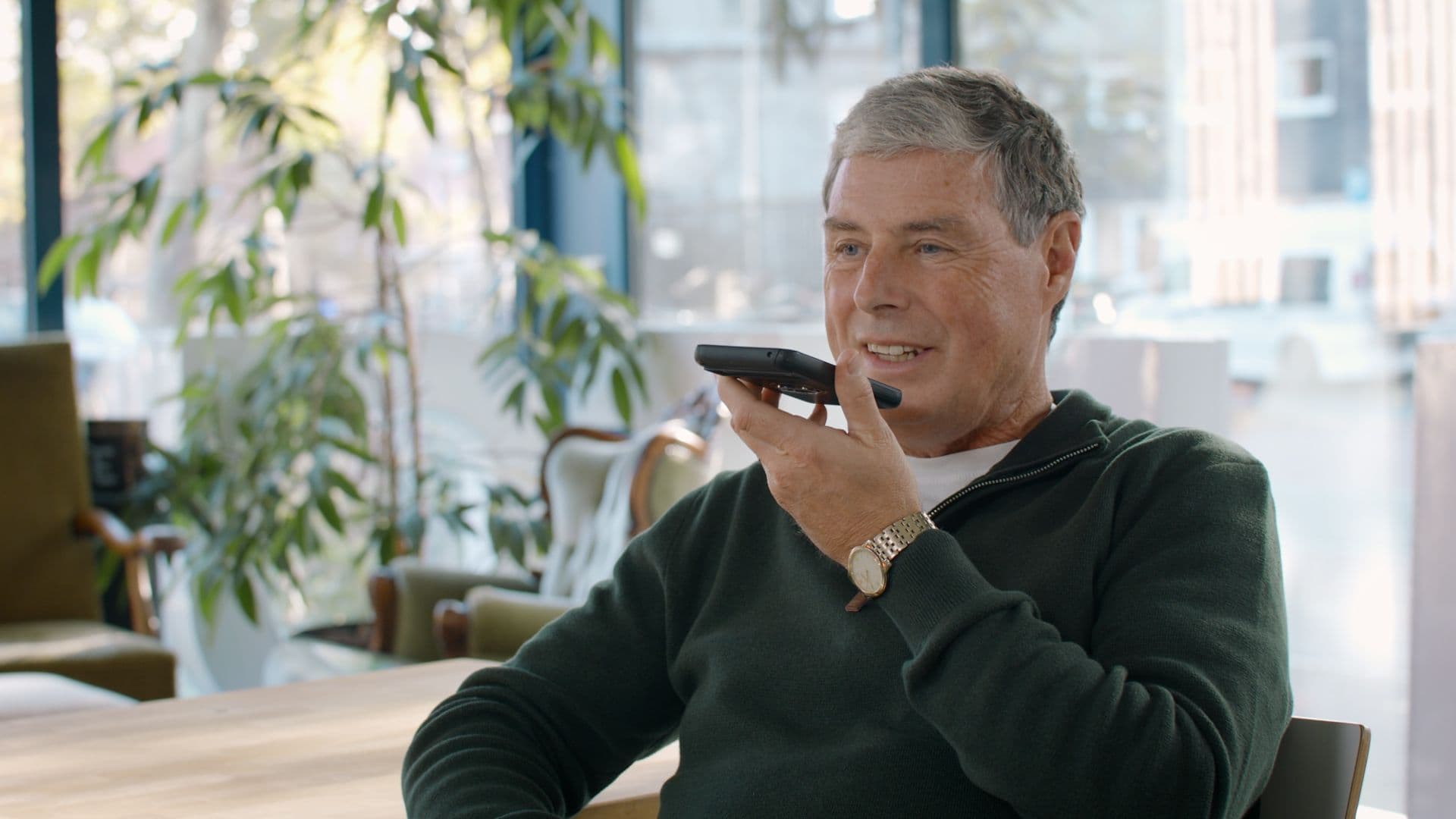 An older white man speaking into a smartphone. He is sitting in a chair with a desk and plant behind him.