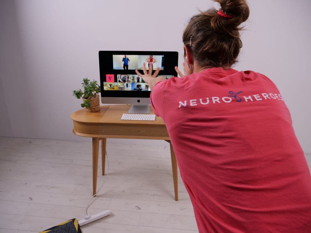 A woman in a red Neuro Heroes t-shirt stretching her arms toward a computer monitor. There is an exercise class on the monitor with multiple people in the call.