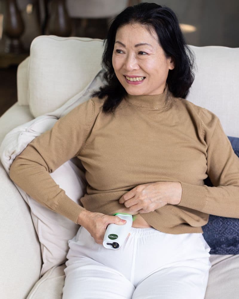 Asian woman holds a white Symbyx device to her bare stomach. Her other hand holds her jumper up.