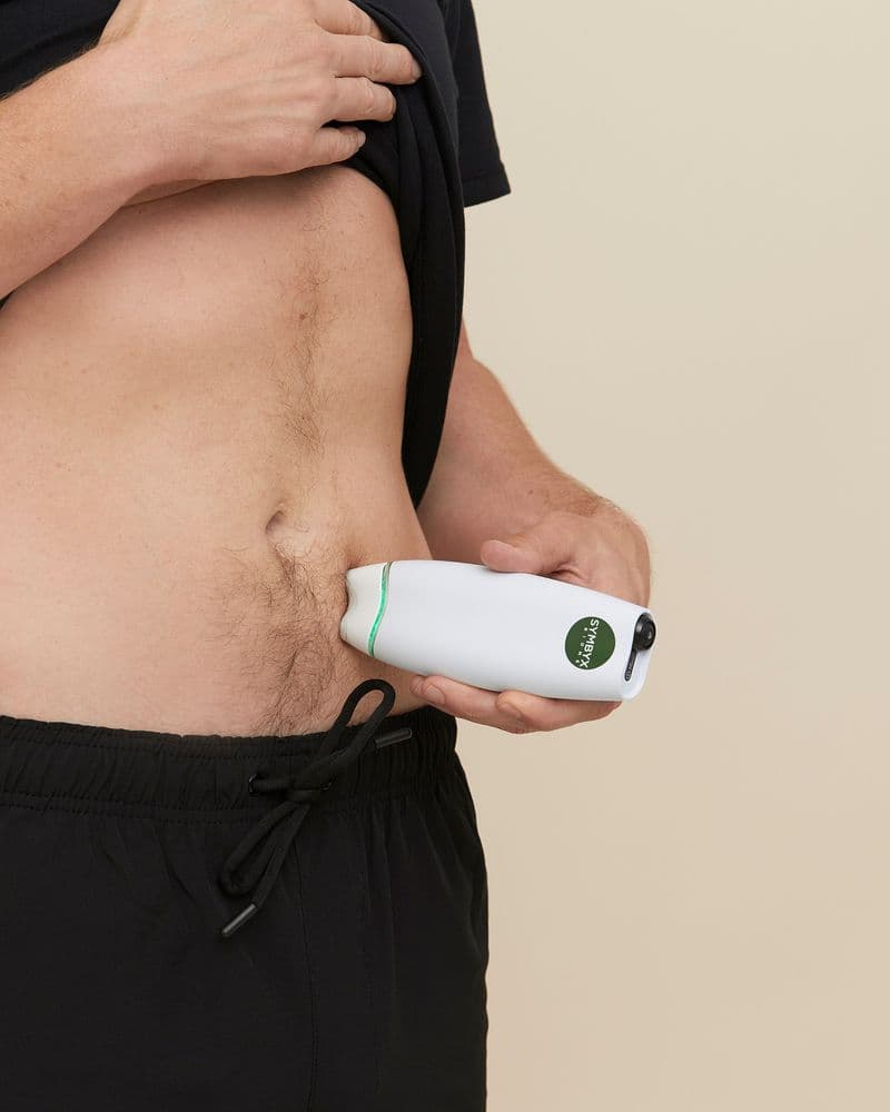White man holds a white Symbyx device to his bare stomach. His other hand holds a black T-shirt up.