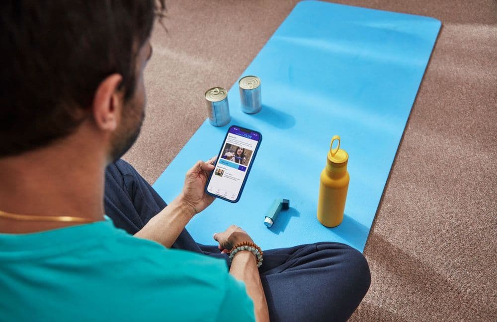A photo of a man sitting on a yoga mat. There are two tin cans, a water bottle and an inhaler next to him. He is holding a phone with the We Are Undefeatable app visable on screen.