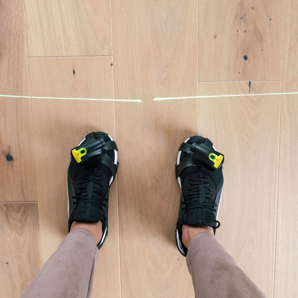 A photo of someone's trainers. The Path Finder device laser is strapped on each trainer. The laser shines on the ground.
