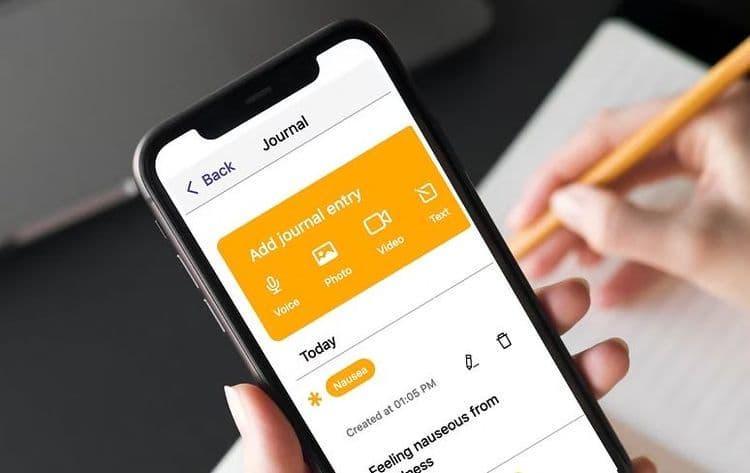 A white hand holding an iPhone with the My Moves Matter app on screen. The app is on the journal entry page, while the person's other hand is writing on a sheet of lined paper with a pencil.