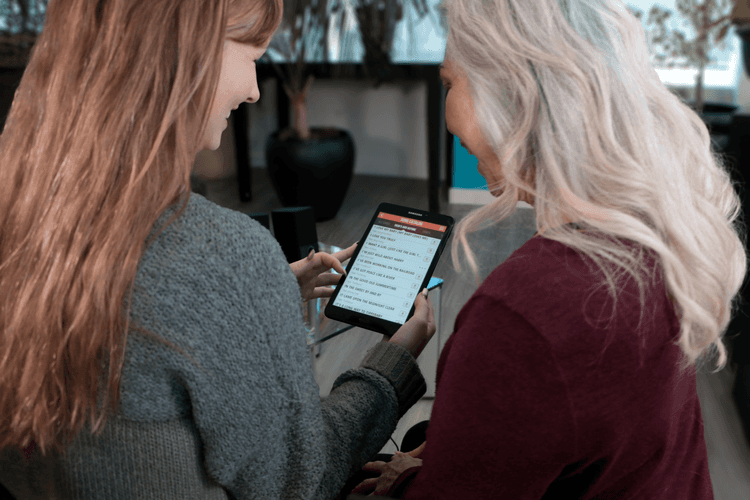 An image of a woman holding a tablet showing a list of songs on the SingFit app. There is an older woman next to her also looking at the tablet. 