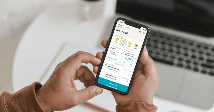 A photo of two hands holding an iPhone in front of a laptop. The phone screen shows the My Day tab in the StrivePD app.
