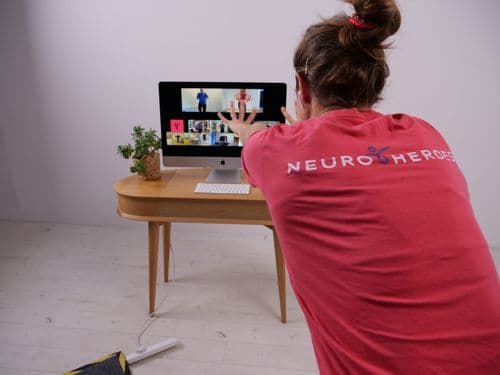 A woman in a red Neuro Heroes t-shirt stretching her arms toward a computer monitor. There is an exercise class on the monitor with multiple people in the call.