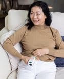 Asian woman holds a white Symbyx device to her bare stomach. Her other hand holds her jumper up.