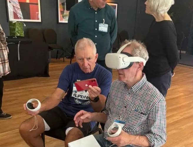 A photo of Keith Wilson holding one VR controller and an iPhone and talking to another man. The other man is wearing a VR headset and holding the other controller. Both are sat down.