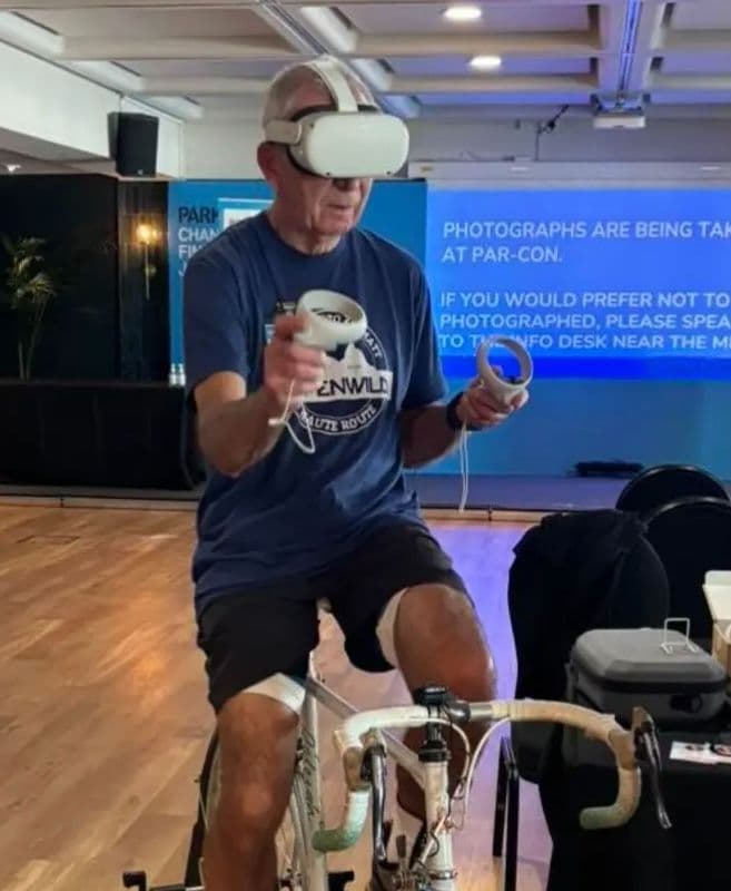 A photo of Keith Wilson wearing a VR headset, holding controllers and sitting on a bike.