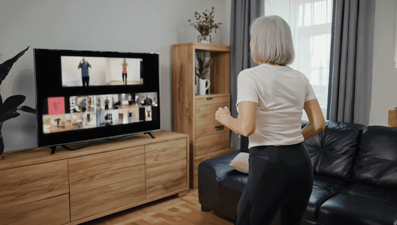 A lady with a grey bob exercising in her living room. She is facing toward a TV where you can see a large group of people working out over video call.