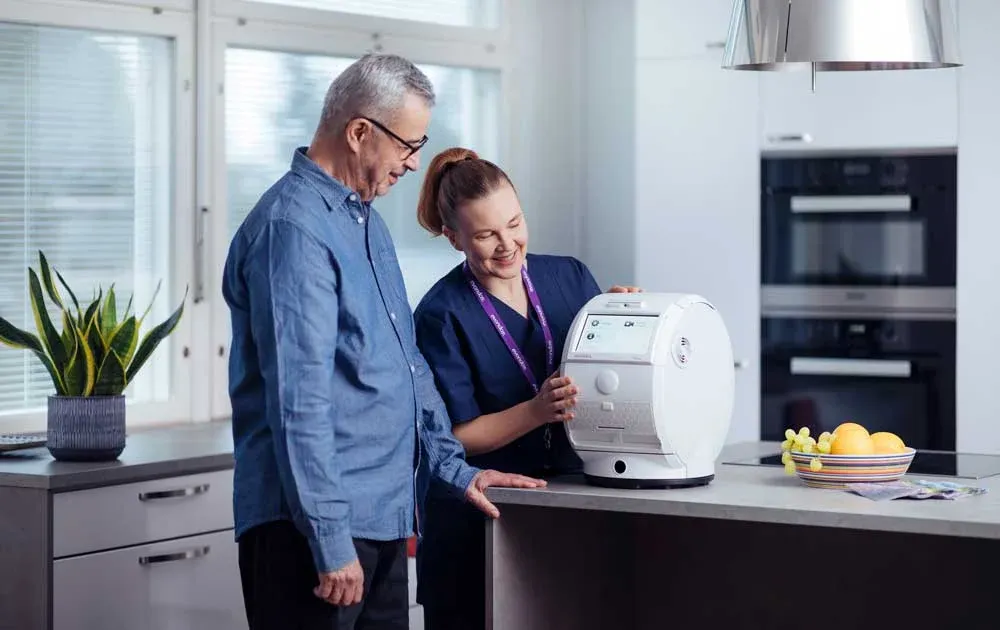 Photo of a carer showing a tall, elderly white man how to use the Evondos robot while in the kitchen.
