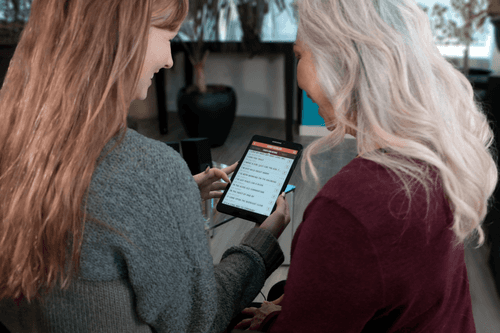 An image of a woman holding a tablet showing a list of songs on the SingFit app. There is an older woman next to her also looking at the tablet.